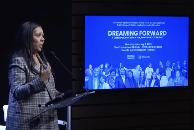 A woman in a plaid blazer speaks at a podium beside a screen promoting the Dream Keeper event hosted by the Human Rights Commission on February 9, 2023.